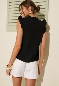 Blusa nera con texture e maniche a volant, abbinata a pantaloni corti bianchi. Include accessori come un bracciale d'oro al polso.