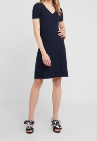Abito a maglia blu navy con scollo a V e maniche corte, caratterizzato da un motivo a coste testurizzato. Indossato con sandali neri e decorazioni a forma di fiocco a fantasia.