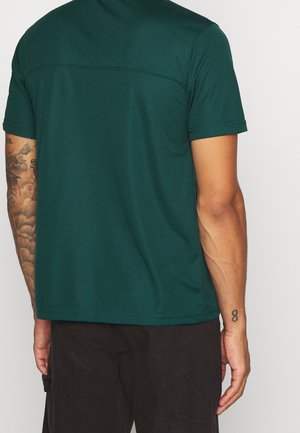 Uomo con camicia verde scuro a maniche corte e pantaloni neri, mostra tatuaggi sul braccio sinistro e un tatuaggio del numero 8 sul polso destro, in piedi contro una parete bianca.