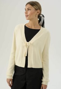 Cardigan color crema in materiale morbido, con scollatura a V, polsini a costine e dettaglio con fiocco sul davanti. Abbinato a un top nero e pantaloni.