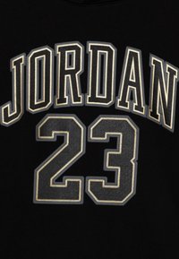 Černá tkanina s vyšitým nápisem "JORDAN" v tučném bílém a šedém písmu nad číslem "23" ve podobném vyšitým stylu, které vykazuje texturovaný vzhled.