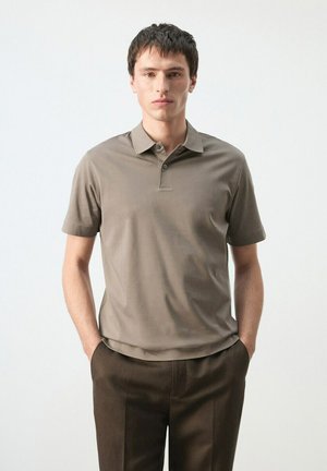 Joven de pie con las manos en los bolsillos, vistiendo una camisa tipo polo de manga corta color topo y pantalones marrón oscuro, frente a un fondo liso.