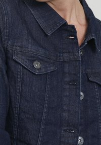 Mörk denimjacka med klassisk krage, två bröstfickor med klaff och knappar med silvrig ton, som visar en texturerad tygyta.