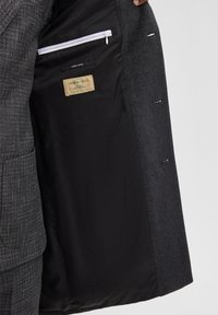 Grijze wollen blazer met een zwarte satijnen voering. Beschikt over een ritszak en een label met de tekst "GREENWOOL." Geverfde buitenstof met patroon.