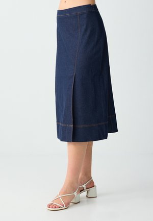 Kobieta ubrana w ciemnoniebieską jeansową spódnicę midi z rozcięciem z przodu oraz białe sandały na słupku z paskami, stojąca na jednolitym tle.