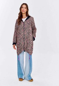 Žena v dlouhém leopardím kabátu s růžovými akcenty, světle modrých širokých džínách a hnědých platformových botách, stojící na bílém pozadí.