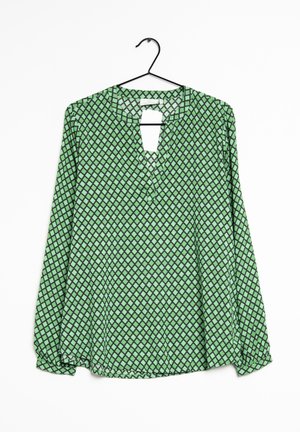 Blouse verte à manches longues avec motif de losanges noirs, col rond avec fente à l'avant, suspendue à un cintre noir sur fond blanc.