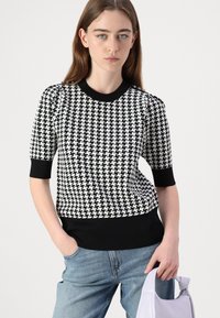 Sweter z czarno-białym wzorem w jodełkę, dziany, z krótkimi rękawami oraz czarnym prążkowanym dekoltem i dolnym wykończeniem, noszony z jasnoniebieskim dżinsami.