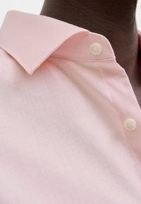 Ροζ πουκάμισο με λεπτή υφή, καλοσχηματισμένο γιακά και ένα μόνο λευκό κουμπί στον λαιμό.