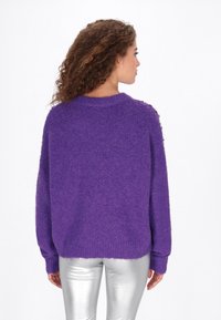 Paarse gebreide trui met een stevige textuur en een ronde halslijn, versierd met decoratieve knopen op de schouder, gecombineerd met glanzende zilveren broek.