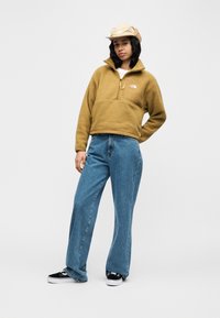 Νεαρή γυναίκα με μπεζ φλις μπουφάν, καπέλο που ταιριάζει, φαρδιά μπλε τζιν και ασπρόμαυρα αθλητικά παπούτσια, στέκεται σε λευκό φόντο.