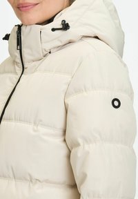 Vrouw gekleed in een lichtbeige gewatteerde winterjas met een capuchon en zwarte ritssluiting, gedeeltelijk gezicht zichtbaar, close-up van het bovenlichaam.