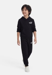 Παιδί που περπατά φορώντας μαύρο φούτερ με κουκούλα Puma και φόρμες, με λευκά παπούτσια slip-on, κοιτάζει προς τα πλάγια και χαμογελά.