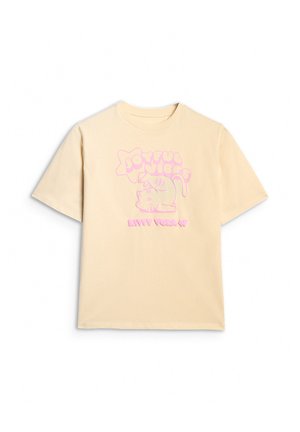 Młoda dziewczyna ubrana w beżowy, oversize'owy t-shirt z nadrukiem, szare rozszerzane spodnie, szare trampki oraz małą różową torebkę na ramię na jednolitym tle.
