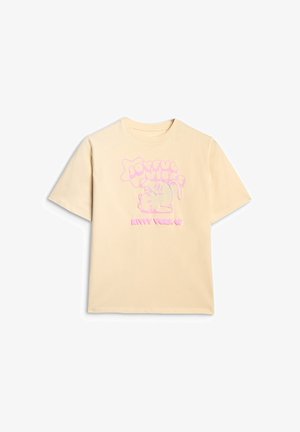 Młoda dziewczyna ubrana w beżowy, oversize'owy t-shirt z nadrukiem, szare rozszerzane spodnie, szare trampki oraz małą różową torebkę na ramię na jednolitym tle.