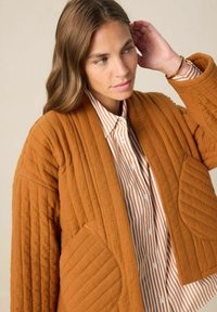 Veste matelassée en orange brûlé avec une texture côtelée et de grandes poches, portée par-dessus une chemise à rayures blanches et rouille.