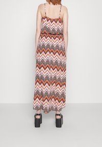 Vrouw die een mouwloze maxi-jurk draagt met rood-witte- en zwarte zigzagpatronen, gecombineerd met zwarte platformsandalen, staand met de rug naar de kijker.