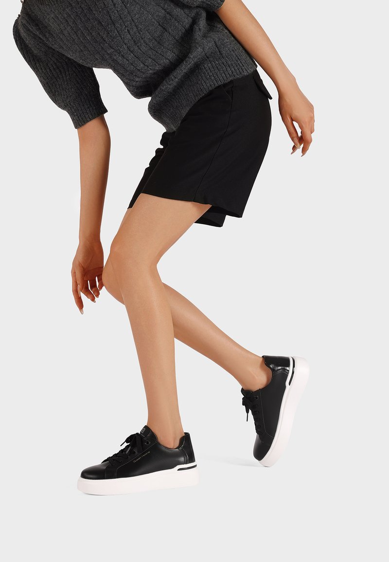 Persona che indossa sneakers nere con suole bianche e pantaloni corti neri, inclinata in avanti contro uno sfondo chiaro e semplice.