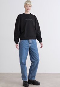 Svart hettegenser med ribbet mannskapskrage, kombinert med løse lyseblå jeans. Sko med kraftige svarte såler. Enkelt design generelt.