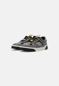 Black and gray sneakers with a textured finish, padded tongue, yellow laces, and a chunky white sole. Features angular design elements.