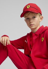 Raudonas sportinis kostiumas, sudarantis užtrauktuką turintį džemperį ir kelnes su „Ferrari“ prekės ženklu. Pridedama deranti raudona kepurė su logotipu. Minkšta tekstūra.