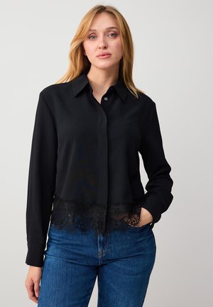 Kobieta o blond włosach, ubrana w czarną bluzkę z długim rękawem z koronkowym wykończeniem oraz niebieskie dżinsy, stojąca z jedną ręką w kieszeni na szarym tle.