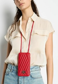 Custodia per telefono rossa trapuntata con cuciture diagonali, caratterizzata da un logo dorato. Sospesa da una tracolla coordinata, indossata sopra una camicia leggera e setosa.