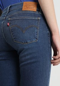 Acercamiento de una persona que lleva jeans Levi's azules, mostrando el bolsillo trasero, la etiqueta roja de Levi's y el parche de cuero de la marca.