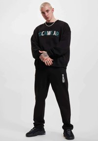 Mustassa hupparissa on rohkea turkoosi "ROCWEAR" -teksti, ja se on yhdistetty mustiin verkkareihin, joissa on pieni logo. Asusteena mustat lenkkarit.