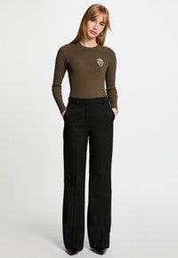Femme se tenant debout les mains dans les poches, portant un haut long à manches ribblé olive avec un blason et un pantalon noir taille haute à jambes larges, ainsi que des chaussures noires.