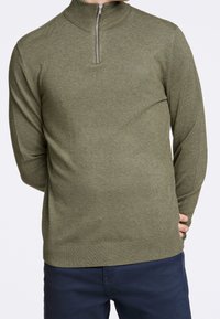 Pull en tricot vert avec un col haut et une fermeture éclair mi-hauteur. Présente des manches longues et une texture lisse et douce. Porté avec un pantalon bleu marine.