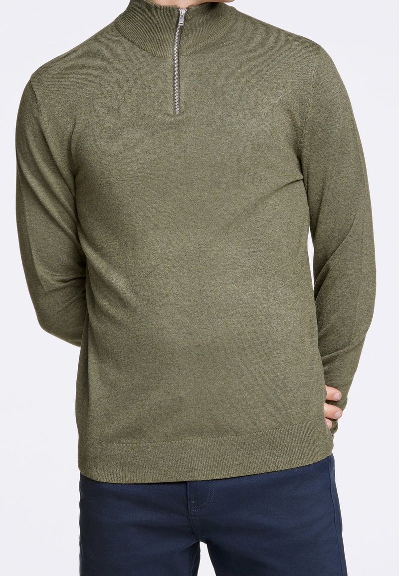Pull en tricot vert avec un col haut et une fermeture éclair mi-hauteur. Présente des manches longues et une texture lisse et douce. Porté avec un pantalon bleu marine.