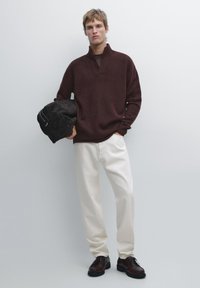 Brązowy sweter z dzianiny z kołnierzem, kremowe szerokie spodnie i ciemne buty. Modelka trzyma ciemną torbę na jednolitym tle.