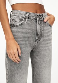 Femme portant un jean noir délavé taille haute, une main dans la poche et l'autre reposant sur son côté, avec un top blanc court.