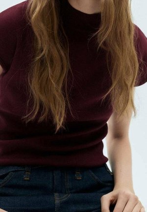 Młoda kobieta z długimi falującymi jasnobrązowymi włosami, ubrana w bordową bluzkę z krótkim rękawem i ciemnoniebieskie jeansy, siedząca z jedną ręką opartą na kolanie.