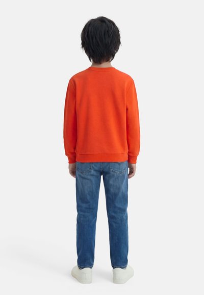 Enfant aux cheveux courts et foncés portant un sweat-shirt orange, un jean bleu et des baskets blanches, debout de dos sur un fond blanc.