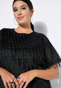 Abito nero con un design a frange testurizzate. Maniche corte, collo tondo e inserti in tessuto trasparente. Più anelli sulle mani.