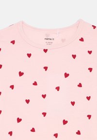 Light pink T-shirt featuring a pattern of small red hearts, made of soft cotton fabric with a round neckline and short sleeves.