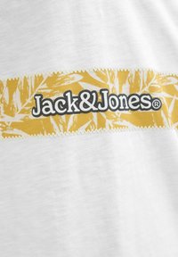 Hvid T-shirt med en gul stripe med bladmønster og "Jack&Jones" logo i fed sort skrift centreret på striben.