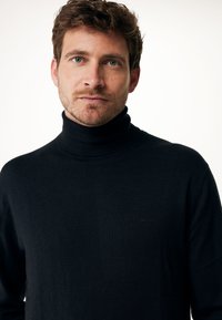 Svart polotröja i en mjuk, texturerad stickning. Har en figurnära design med ribbat krage. Minimal branding synlig på bröstet.