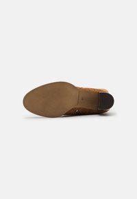 Scarpa in suede marrone con tacco in pelle liscia e suola testurizzata. Presenta un'apertura sul retro e cinturini decorativi sui lati.