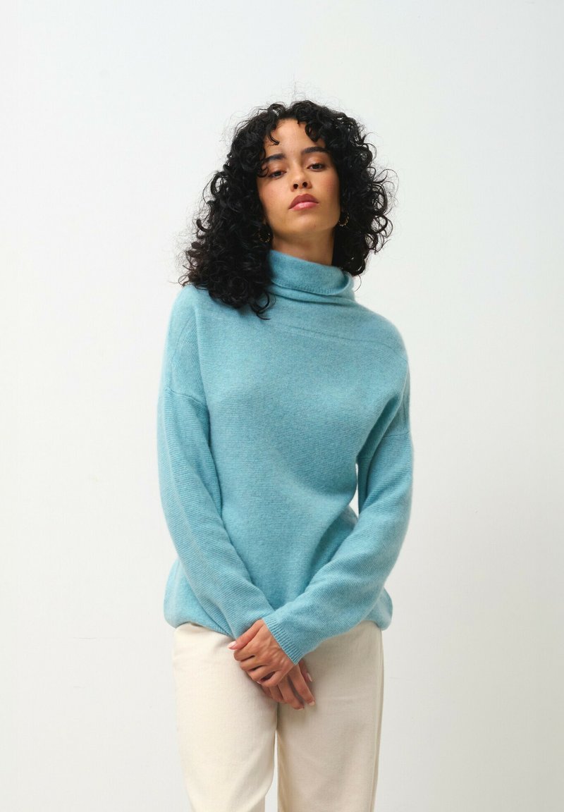 Hellblauer Rollkragenpullover aus weichem Material, mit langen Ärmeln und lässigem Schnitt, kombiniert mit cremefarbenen Hosen.