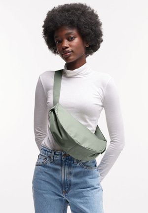 Giovane donna con afro che indossa un dolcevita bianco, jeans blu e una borsa verde a tracolla, in piedi davanti a uno sfondo bianco.