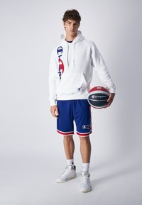 Sudadera de felpa blanca con un gran logo, combinada con pantalones cortos de malla azules con ribetes rojos. El modelo sostiene una pelota de baloncesto negra y roja. Zapatillas blancas.