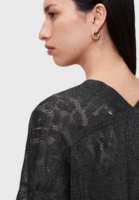 Schwarzes gestricktes Oberteil mit einem strukturierten, offenen Gewebemuster. Der Rücken hat einen V-Ausschnitt und präsentiert das Seitenprofil des Models sowie geometrische Ohrringe.