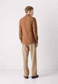 Camicia marrone a maniche lunghe, texture liscia, polsino abbottonato, abbinata a pantaloni beige, slim fit, scarpe nere, sfondo uniforme.