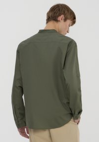 Camicia a maniche lunghe verde oliva con colletto semplice e polsini abbottonati, caratterizzata da una texture liscia e una vestibilità rilassata.
