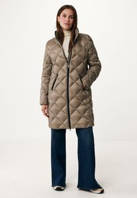 Gequilted lange jas in lichtbruin met ritsluiting, hoge kraag en zijzakken. Gepaard met donkere flared jeans en sneakers.