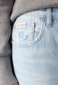 Vaqueros de mezclilla de color azul claro con detalles desgastados en los bolsillos, que presentan una etiqueta roja de "Calvin Klein" y llamativos accesorios en plata. Textura suave.
