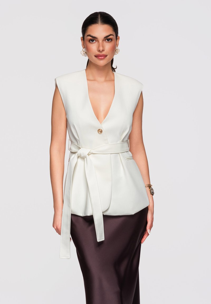 Femme aux cheveux foncés portant un blazer blanc sans manches ceinturé et une jupe en satin foncé, posant devant un fond clair uni.